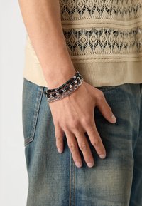Sort og sølv multistrenget armbånd på en hånd, med perler og kædelinks, sat mod et strikket beige plagg. Jeans er synlige.