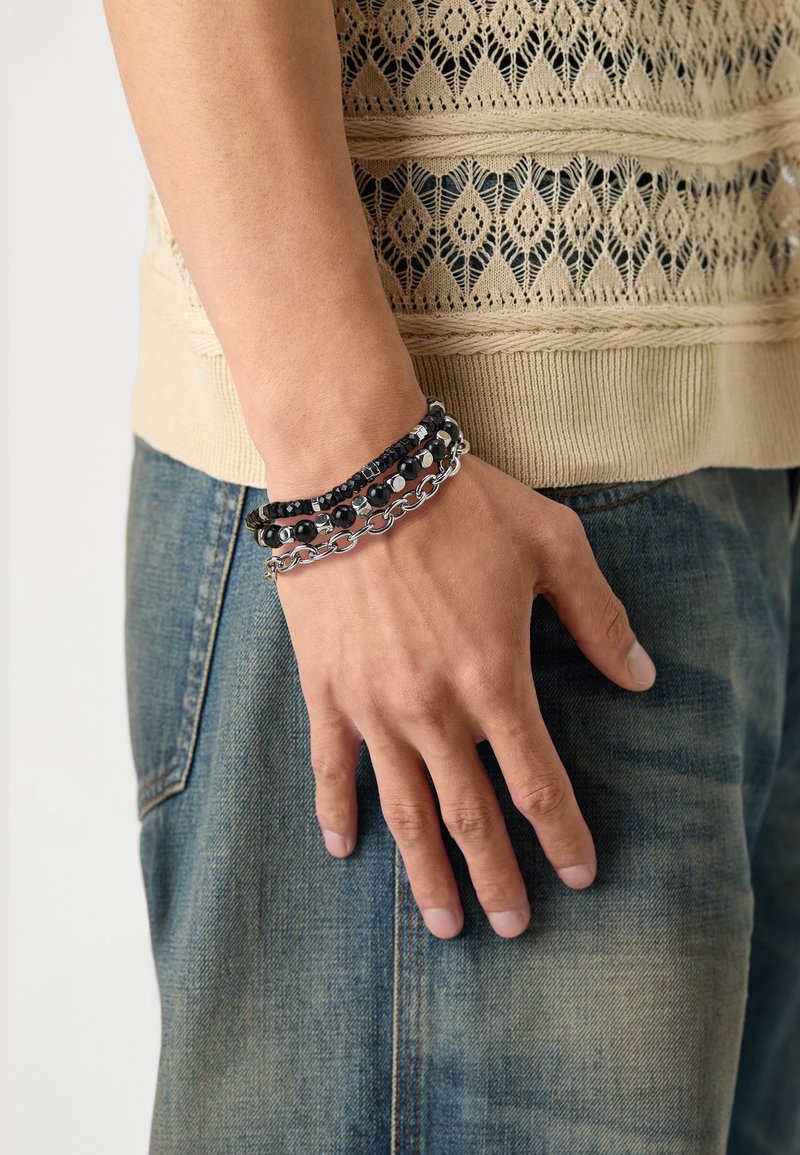Sort og sølv multistrenget armbånd på en hånd, med perler og kædelinks, sat mod et strikket beige plagg. Jeans er synlige.