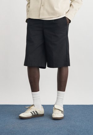 Bărbat purtând pantaloni scurți negri până la genunchi, șosete albe până la gleznă și adidași albi cu dungi negre, stând pe un covor albastru.