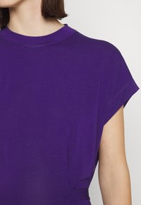 Fialový top s krátkým rukávem a kulatým výstřihem, vyrobený z hladké, lehké tkaniny s jemnými bočními detaily.