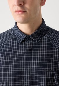 Ναυτικό πουκάμισο με καρό σχέδιο, με λευκό πλέγμα σε αντίθεση, διαθέτει γιακά και διακριτική λεπτομέρεια λογότυπου στο στήθος. Υφασμάτινη υφή.
