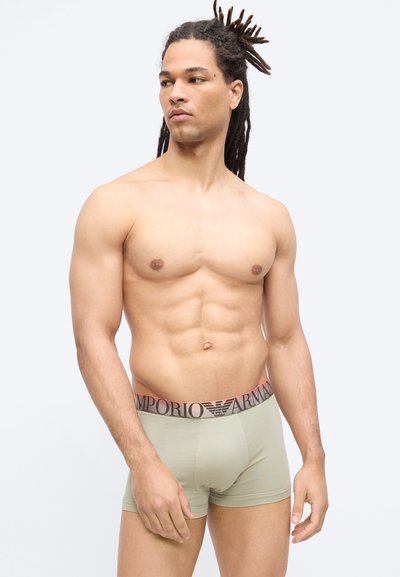 Ανδρας χωρίς μπλούζα με dreadlocks, φορώντας ανοικτό πράσινο εσώρουχο Emporio Armani, κοιτάζει στο πλάι μπροστά σε απλό λευκό φόντο.