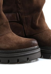 Gros plan sur des bottes en daim marron avec des semelles épaisses en caoutchouc noir à crampons et un petit détail de sangle en cuir près de la cheville.