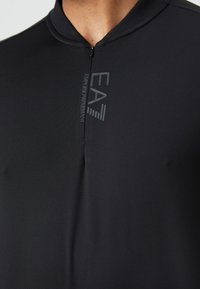 Camisola atlética preta com uma superfície texturizada, fecho de meio zíper, e o logo EA7 no peito. Apresenta um design minimalista e um colarinho alto.