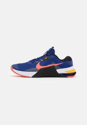 Niebieski sportowy tenis z pomarańczowym logo, czarno-białą podeszwą, żółtymi akcentami i siateczkową cholewką, pokazany z boku na białym tle.