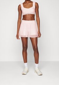 Světle růžový sportovní podprsenka se čtvercovým výstřihem a nabíranými detaily, spárovaná s odpovídající průhlednou sukní. Měkká, lehká tkanina s elastickým pasem.