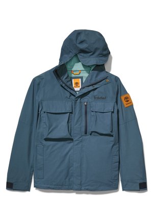 Veste Timberland bleue avec capuche, plusieurs poches avant, fermetures à glissière et boutons, et écusson logo Timberland sur la manche gauche.