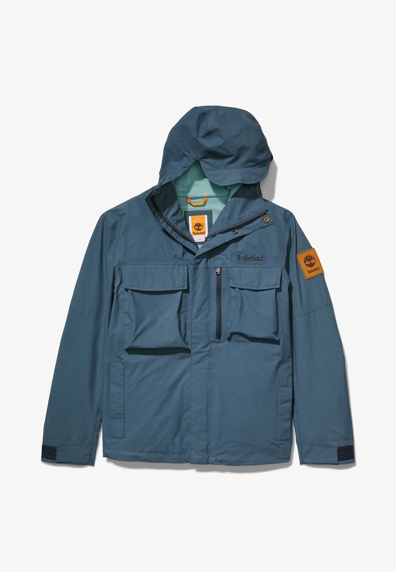Veste Timberland bleue avec capuche, plusieurs poches avant, fermetures à glissière et boutons, et écusson logo Timberland sur la manche gauche.