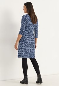 Frau steht mit dem Rücken zur Kamera, trägt ein knielanges blaues Blumenkleid, schwarze Leggings und schwarze Stiefeletten vor einem weißen Hintergrund.
