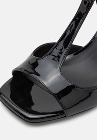 Sandale noire en cuir verni à bout ouvert avec une bride en T et un bout carré, montrant des coutures détaillées et une finition brillante sur un fond clair.