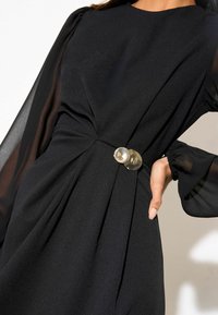 Sort kjole med tætsiddende liv, flydende gennemsigtige ærmer og en vridningsdetalje ved taljen fremhævet af en skinnende rund knap.