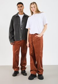 Zwei Modelle stehen nebeneinander. Eine trägt eine schwarze, übergroße Jacke aus Kunstleder über einem weißen T-Shirt, und die andere trägt ein kurzes weißes T-Shirt und rostfarbene Cordhosen. Beide haben klobige schwarze Schuhe an.