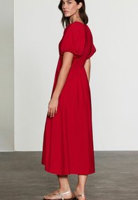 Robe rouge mi-longue avec des manches bouffantes, taille élastique et tissu lisse. Le modèle porte des sandales dorées, se tenant de profil contre un fond neutre.