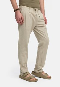 Beige strukturerade byxor med dragkedja i midjan, skräddarsydd passform och sidofickor, i kombination med ljusbruna sandaler.
