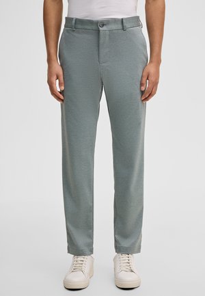 Mann trägt hellgraue gerade geschnittene Hose und weiße Turnschuhe, steht vor einem einfarbigen Hintergrund.