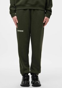 Spodnie dresowe w oliwkowym zielonym kolorze z nadrukiem "MIDTOWN" w białym kolorze. Wykonane z miękkiego materiału, posiadają elastyczne mankiety i luźny krój. W połączeniu z czarnymi sneakersami.
