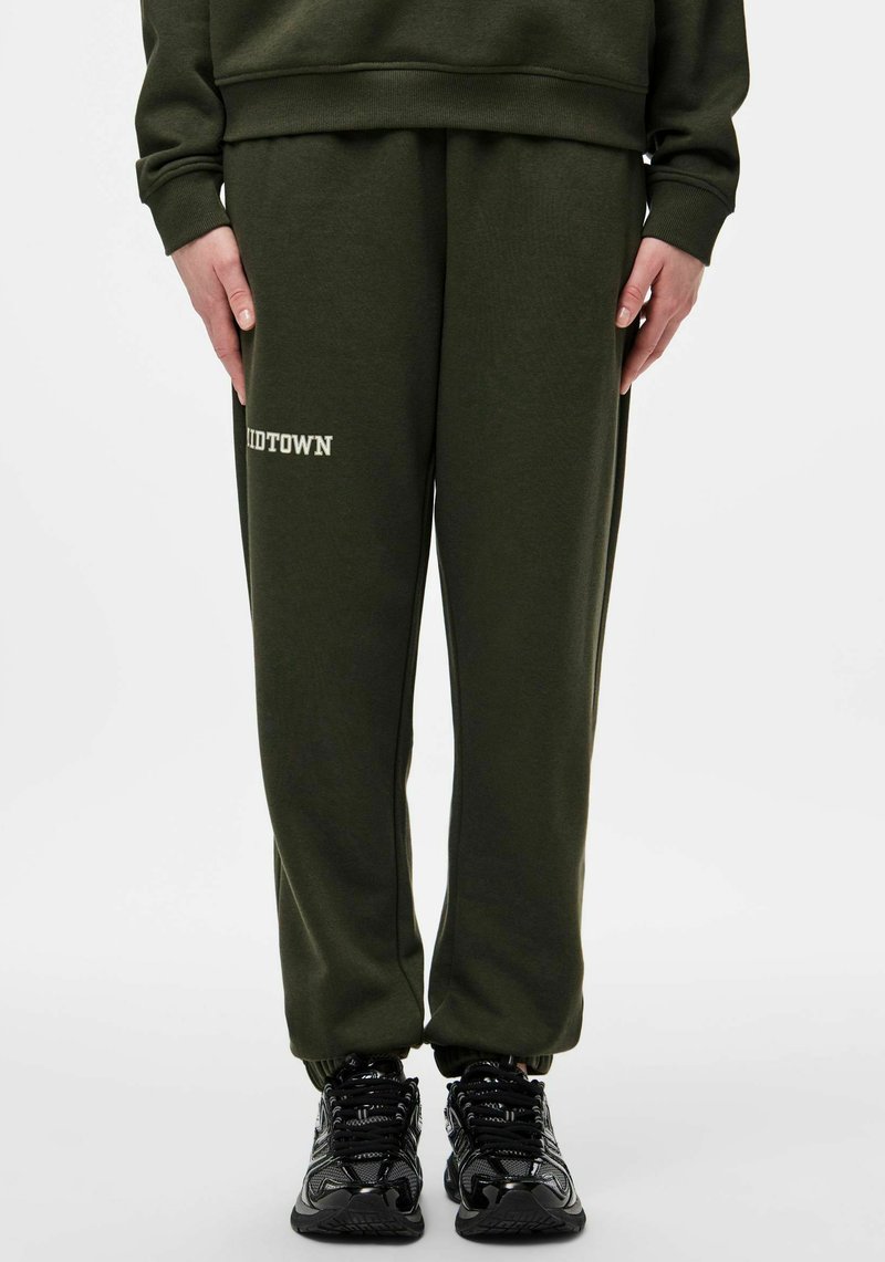 Spodnie dresowe w oliwkowym zielonym kolorze z nadrukiem "MIDTOWN" w białym kolorze. Wykonane z miękkiego materiału, posiadają elastyczne mankiety i luźny krój. W połączeniu z czarnymi sneakersami.