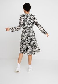 Svart och vit blommig klänning med långa ärmar, snurrad midja och svängd kjol. Bärs med vita sneakers, vilket visar en avslappnad passform och design.