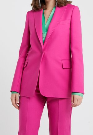 Mujer vestida con un traje sastre de color rosa brillante con pantalones a juego y una blusa verde debajo, complementada con largos pendientes colgantes.