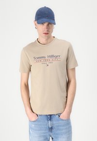 Beige bomull t-skjorte med "Tommy Hilfiger" logo i marineblått og rødt. Båret med lyseblå slitte jeans og en blå caps.