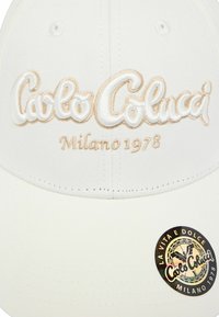 White baseball cap made of fabric with embroidered script "Carlo Colucci Milano 1978" in gold and white, featuring a round logo sticker on the brim.