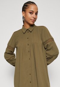 Olive green shirt dress in lightweight fabric, featuring a collared neckline, front button closure, pintuck detailing, and lace accents on sleeves.