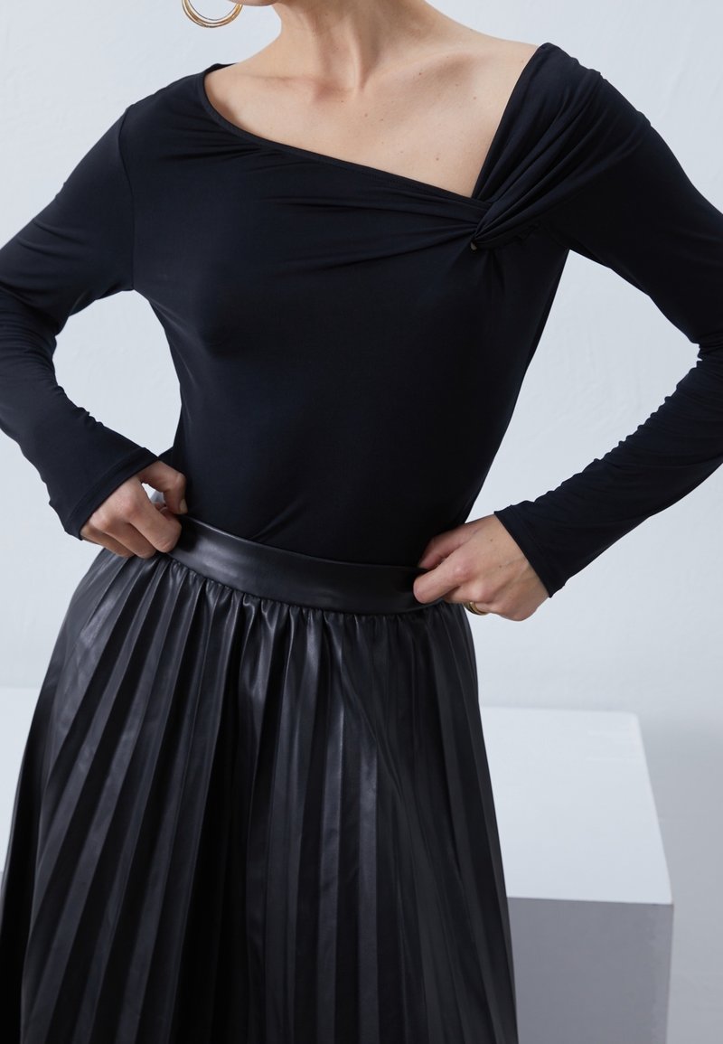 Black long-sleeve top with an asymmetrical neckline and twist detail; paired with a pleated black leather skirt featuring a wide waistband.