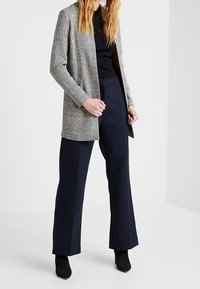 Blazer largo de cuadros gris sobre una blusa negra, combinado con pantalones de pierna ancha en color navy. Botines negros completan el look. Textura de tela suave.