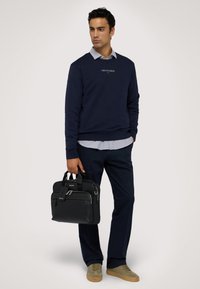 Mörkblå sweatshirt med "TRUSSARDI"-logotyp, ljusblå skjorta med krage under, svart läderväska, mörka byxor och olivgröna skor.