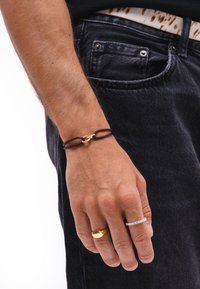 Brown cord bracelet featuring a gold link clasp, worn on a hand with gold and silver rings, against dark denim jeans.