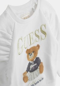 Biely bavlnený sveter s dizajnom zťahovaných rukávov, so zlatým logom "GUESS" a grafikou medveďa v námořníckej mikine a bielej sukni.