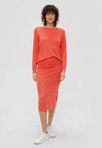Koraalkleurige gebreide trui en midi-jurk met een zigzagpatroon, met een zijsplit. Gecombineerd met witte sneakers voor een casual look.