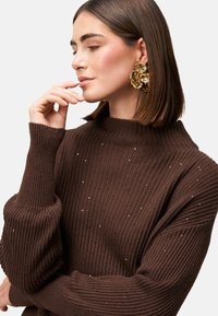 Maglione marrone a coste con un design a collo alto, caratterizzato da sottili decorazioni. Il modello indossa grandi orecchini floreali dorati.