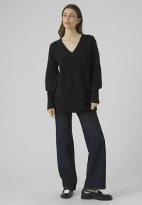 Pullover nero lavorato a maglia con scollatura a V e motivo testurizzato, abbinato a jeans in denim scuro a vita alta e mocassini neri, su uno sfondo neutro.
