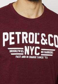 Maroon crewneck-paita, jossa on valkoinen teksti "PETROL & CO NYC," "Brooklyn U.S.," "No Barriers" ja "Fast and in Charge Since '73" rinnassa.