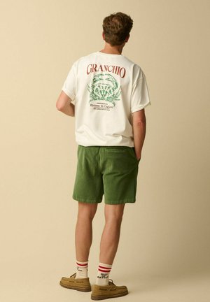 Homme debout de dos, portant un T-shirt blanc avec un graphisme de crabe et du texte, un short vert, des chaussettes rayées et des chaussures marron.