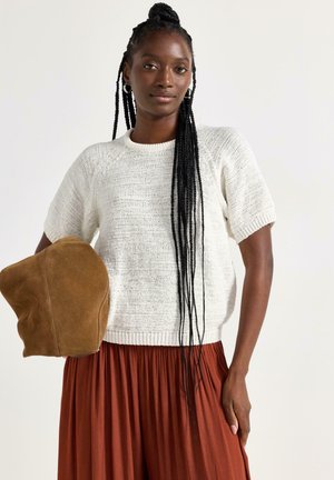 Donna con lunghe trecce che indossa un top bianco a maniche corte in maglia e una gonna plissettata color ruggine, che tiene in mano un cappello di camoscio marrone.
