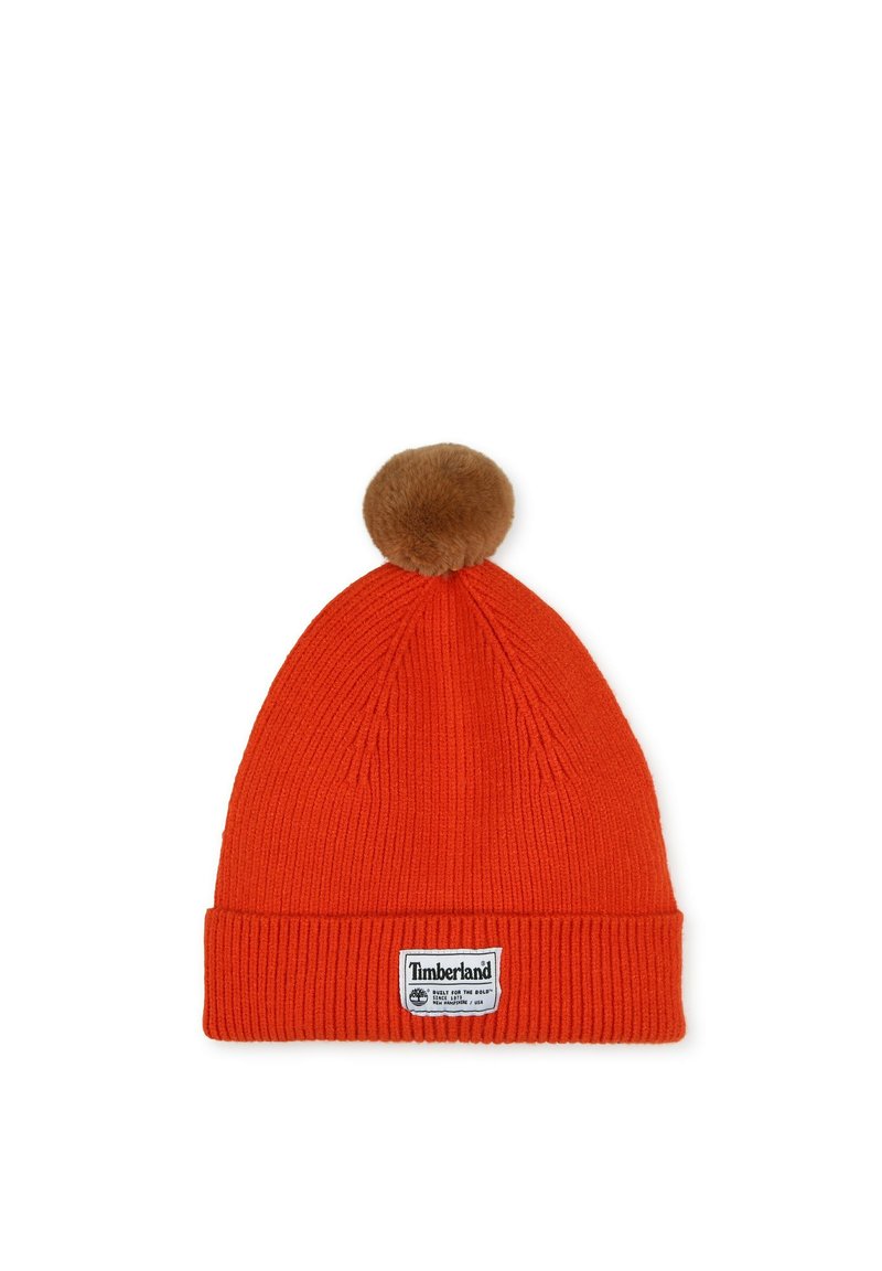 Bonnet en tricot rouge avec une texture côtelée, ornée d'un pompon en fausse fourrure marron sur le dessus et d'un patch logo Timberland blanc sur le revers.