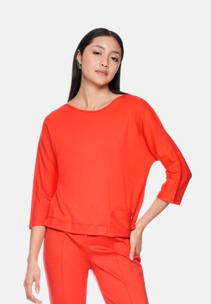 Woman with long dark hair wearing bright red three-quarter sleeve top and matching pants, standing with one hand on hip against white background.