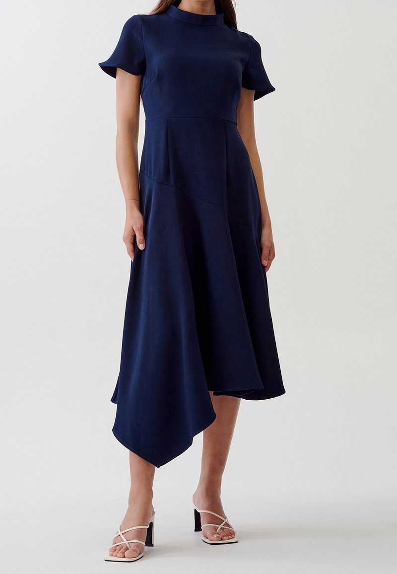 Femme portant une robe midi asymétrique bleu marine à manches courtes avec des sandales à talons blanches, debout devant un fond uni.