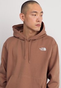 Sudadera marrón hecha de una tela suave, con un bolsillo tipo canguro y cordones. En el frente lleva un pequeño logotipo blanco.