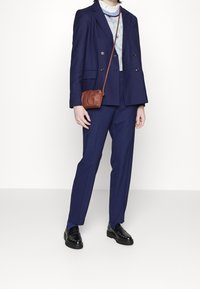 Personne portant un costume bleu marine avec un haut à motifs, des mocassins noirs et un petit sac bandoulière marron sur un fond clair uni.