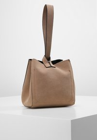 Vaaleanruskea mokkalaukku, jossa on jämäkkä muoto, yläkahva ja etutasku. Minimalistinen muotoilu, sileä pinta ja mustat yksityiskohdat.