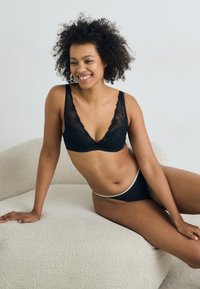 Musta alusvaatesetti, jossa on pitsireunat, sisältäen kolmiomaisen bralette-topin ja siihen sopivat matalavyötäröiset bikinit, esillä teksturoidulla kermaisen vaalealla pinnalla.