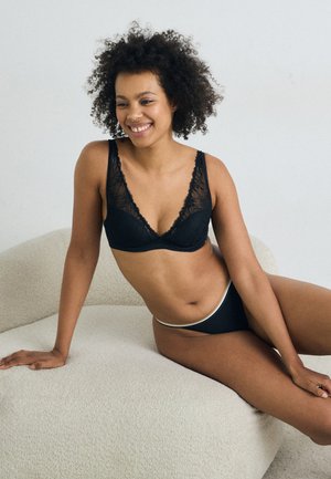 Zwart lingerieset met kantafwerking, bestaande uit een triangle bralette en bijpassende low-rise bikini, tentoongesteld op een gestructureerde crème-kleurige ondergrond.
