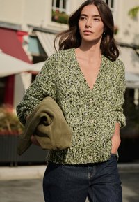 Grøn strikket cardigan med dyb V-udskæring, store knapper og tekstureret mønster. Holder en blød olivengrøn beklædningsgenstand. Mørke jeans fuldender outfittet.