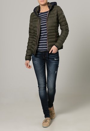 Femme portant une veste matelassée vert foncé, un chemisier rayé bleu marine et blanc, un jean skinny foncé et des mocassins beiges, debout devant un fond uni.