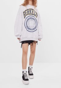 Grå oversized sweatshirt med "BERKELEY" tryckt i gult och blått, med en rund signaturdesign. Bärs med trasiga svarta denimshorts och plattformssneakers.