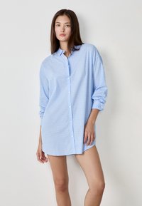Camicia azzurra chiara con bottoni, realizzata in tessuto morbido con un motivo testurizzato, dotata di colletto, maniche lunghe arrotolate fino ai gomiti e orlo arrotondato.