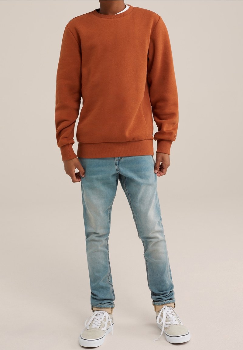 Brun crewneck sweatshirt med ribbede ærmer og bund, parret med lyseblå slidte jeans og beige sneakers med hvide snørebånd.